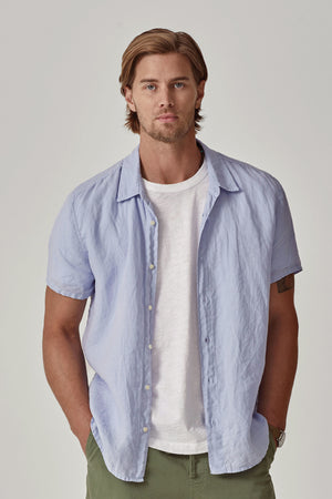 A man with short hair wears the MACKIE LINEN BUTTON-UP SHIRT by Velvet by Graham & Spencer in light blue over a white t-shirt, standing against a plain backdrop. The relaxed fit of his woven linen top enhances the effortlessly casual look.