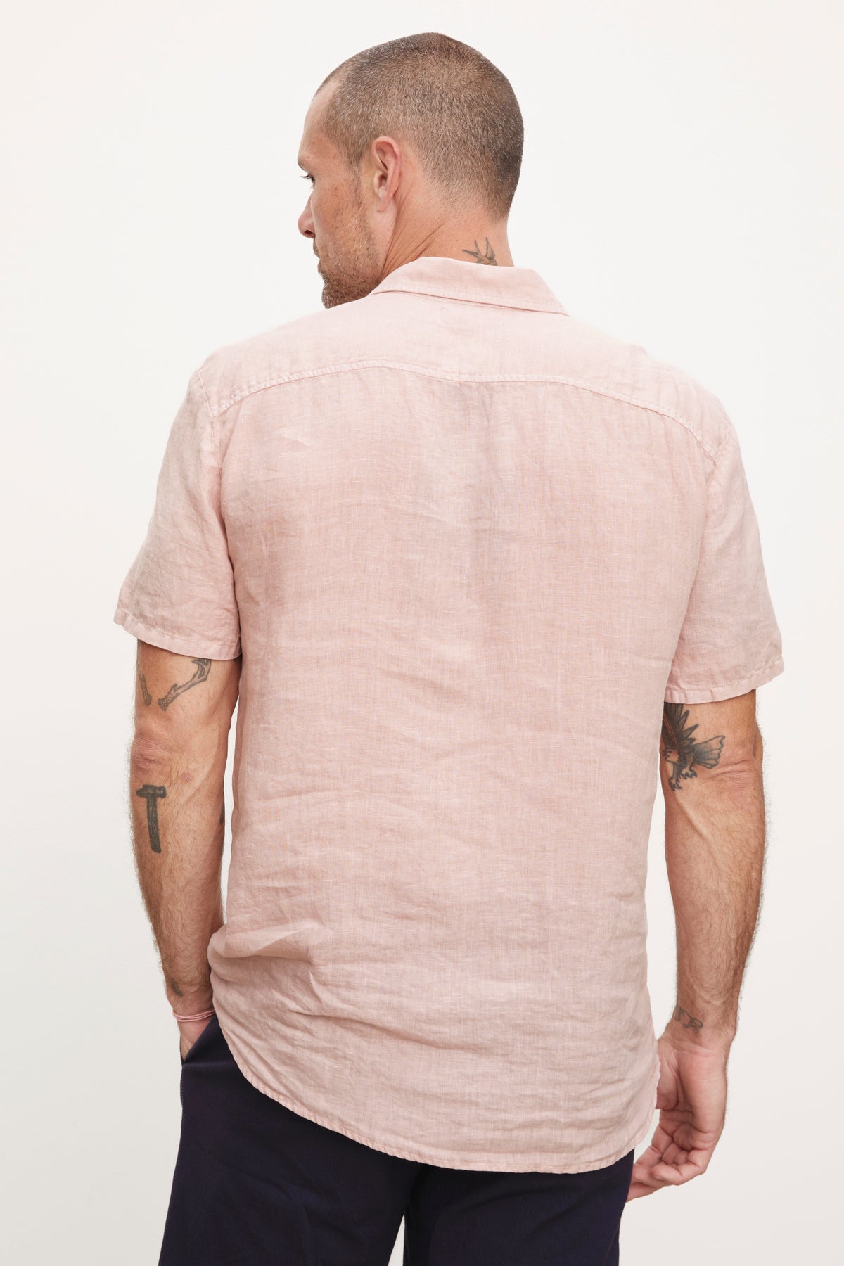   A man viewed from behind, wearing a Velvet by Graham & Spencer MACKIE LINEN BUTTON-UP SHIRT and dark pants, displaying visible tattoos on his arms. 