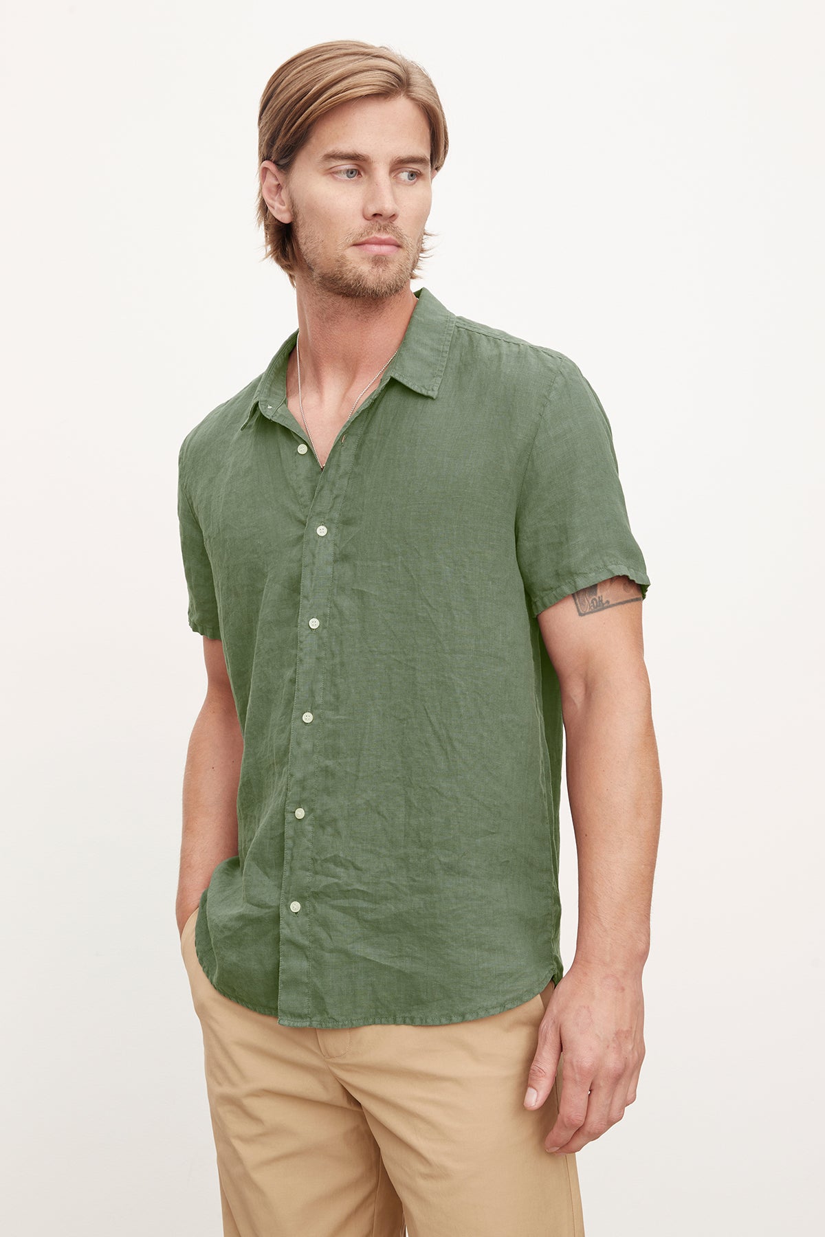   A man with shoulder-length blond hair wearing a Velvet by Graham & Spencer MACKIE LINEN BUTTON-UP SHIRT and beige pants, standing against a light background. 