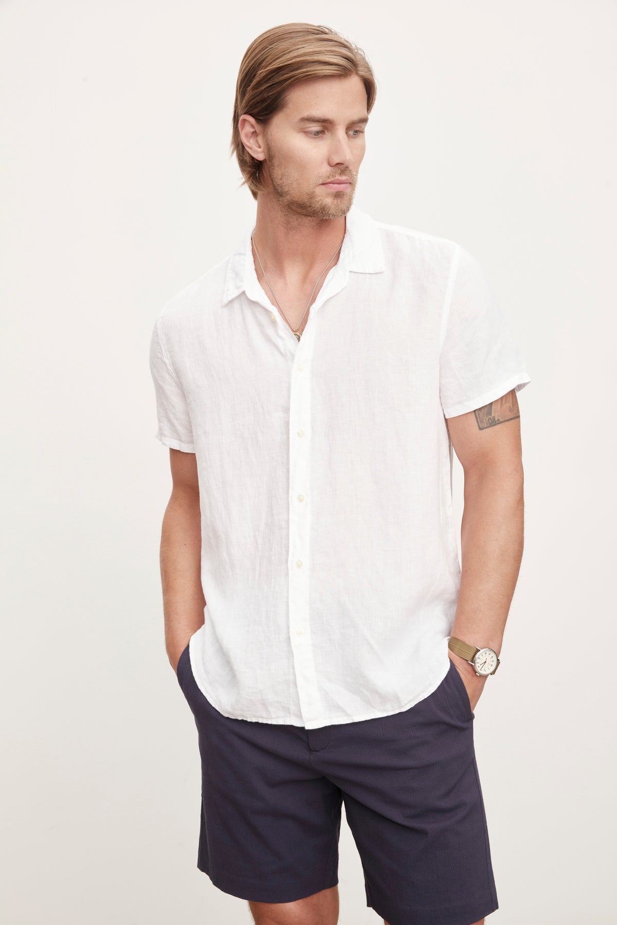   A man in a Velvet by Graham & Spencer MACKIE LINEN BUTTON-UP SHIRT and navy shorts, standing with one hand in his pocket and a watch on his left wrist, epitomizes the relaxed fit of a summer wardrobe. 