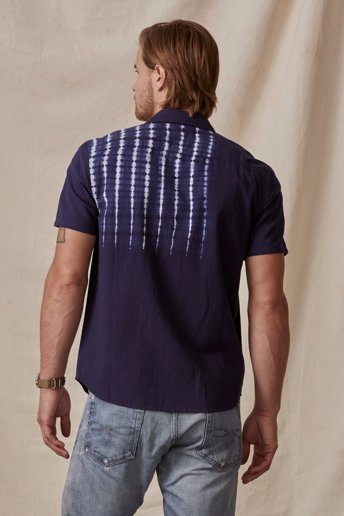   A man with long blonde hair stands with his back facing the camera, wearing a dark purple RAFAEL BUTTON-UP SHIRT by Velvet by Graham & Spencer with a white tie-dye design on the back and light blue jeans. 