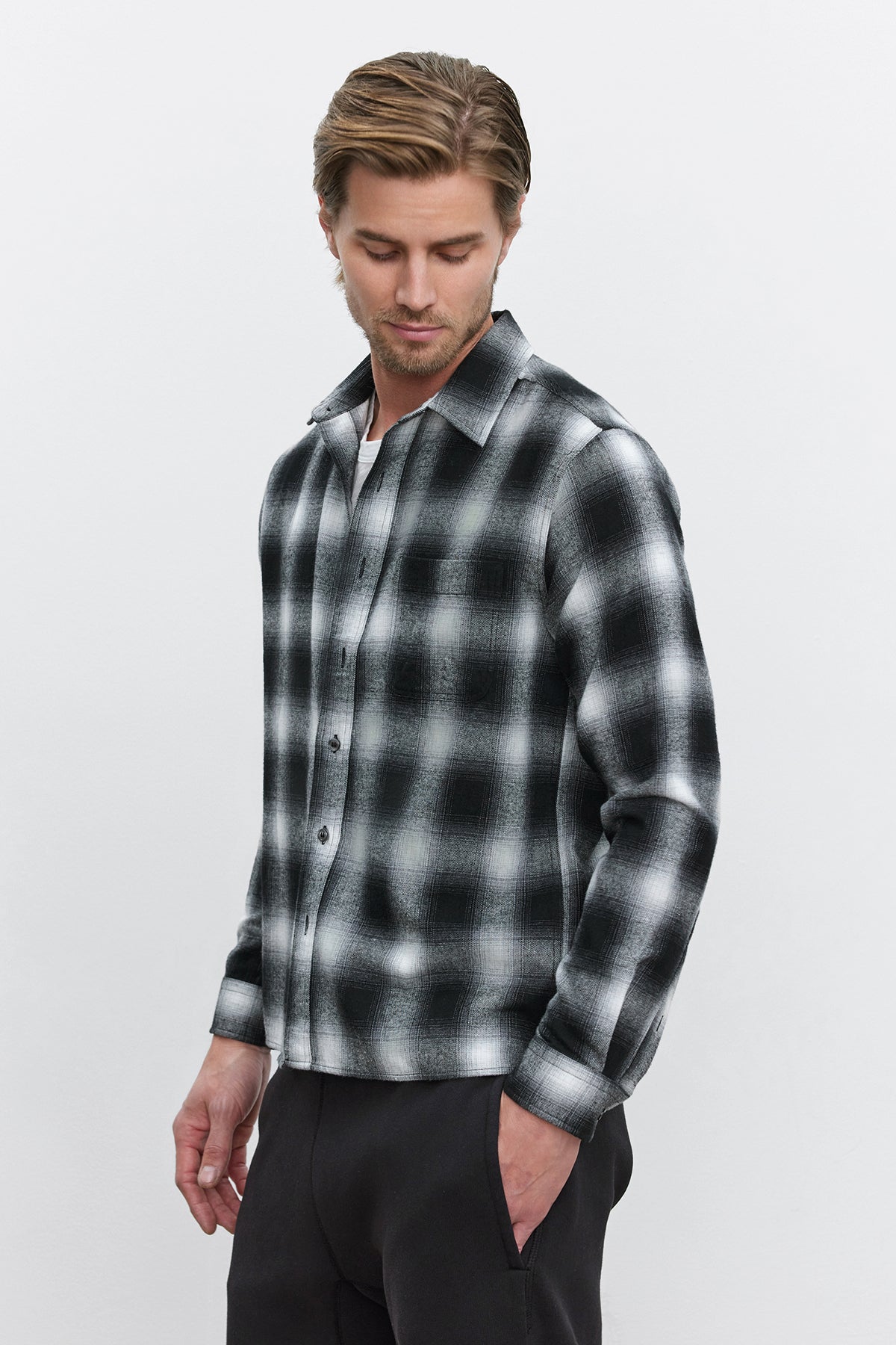   A man is looking down while wearing the STANTON BUTTON-UP SHIRT with a black and white plaid ombre design from Velvet by Graham & Spencer, along with dark pants against a plain background. 