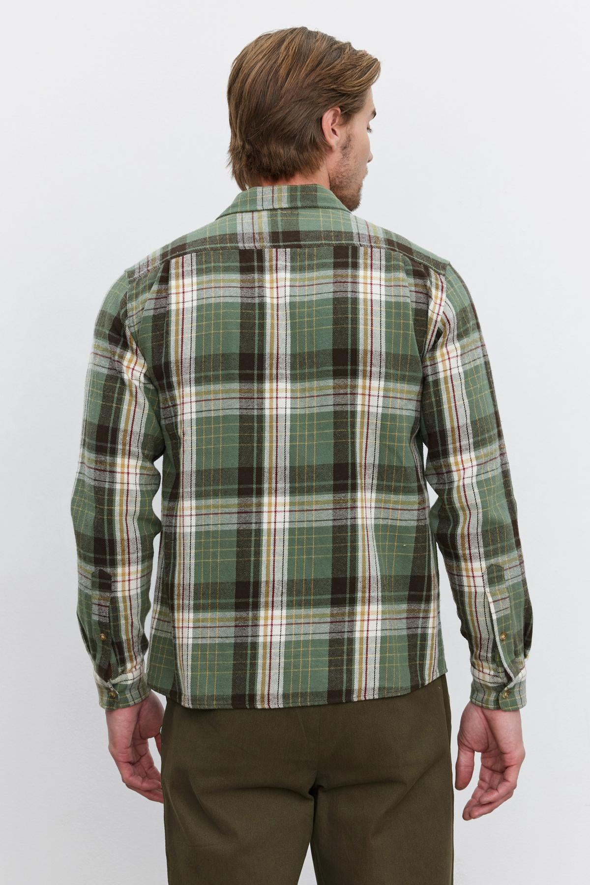   A person with light brown hair is shown from the back, wearing the WAYLON BUTTON-UP SHIRT by Velvet by Graham & Spencer, a green and brown cotton woven flannel plaid shirt with long sleeves, paired with olive green pants. 