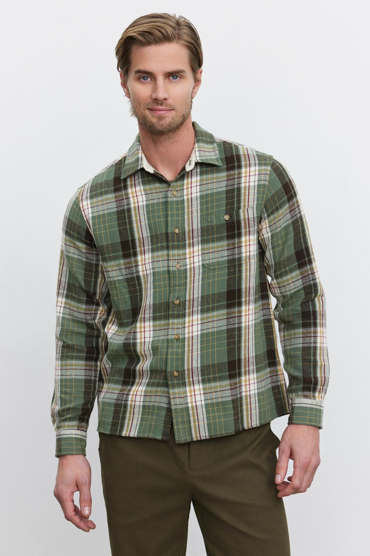   A bearded man wearing the WAYLON BUTTON-UP SHIRT by Velvet by Graham & Spencer, which features a green, brown, and white cotton woven flannel design, stands against a plain background. 