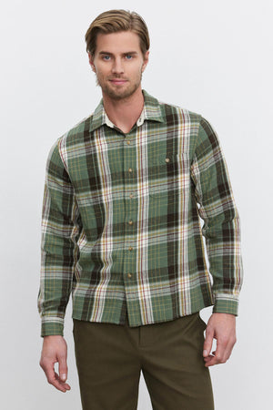 A bearded man wearing the WAYLON BUTTON-UP SHIRT by Velvet by Graham & Spencer, which features a green, brown, and white cotton woven flannel design, stands against a plain background.