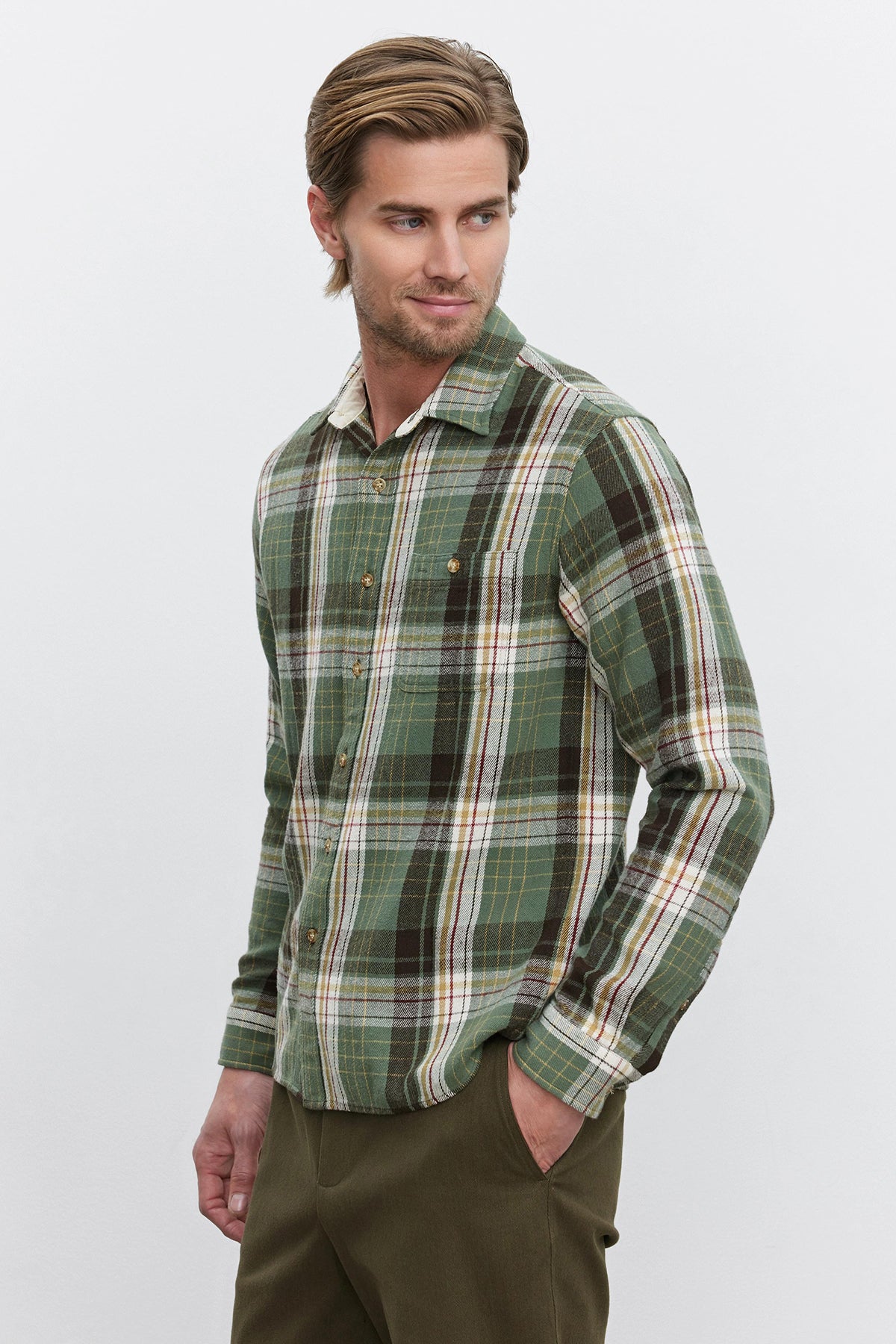   Person wearing a WAYLON BUTTON-UP SHIRT by Velvet by Graham & Spencer, featuring green cotton woven flannel with a chest patch pocket, paired with dark pants. The individual is standing against a plain white background, with one hand in their pocket and looking slightly to the side. 