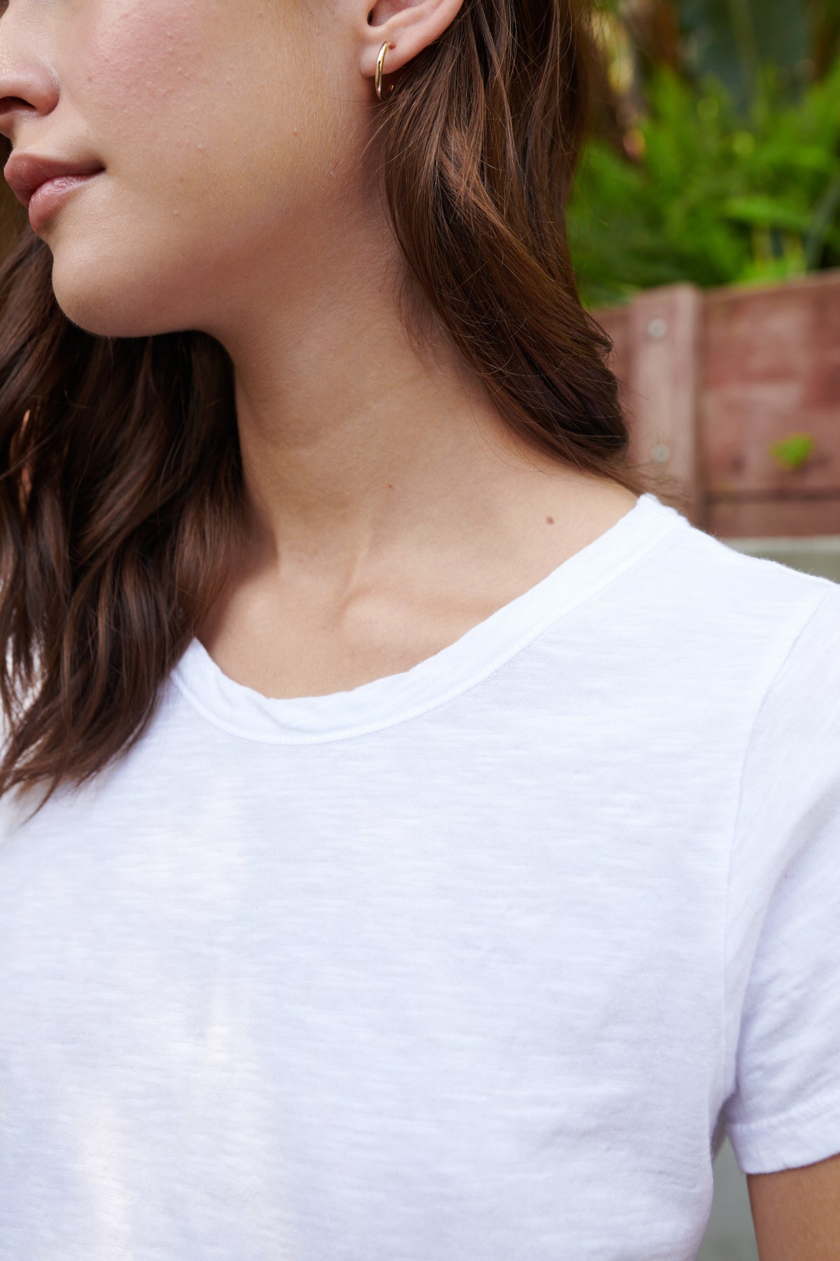   Close-up of a person in a TILLY TEE by Velvet by Graham & Spencer, showing the side of their face and neck with shoulder-length brown hair. The background is slightly blurred with greenery and a wooden structure. 