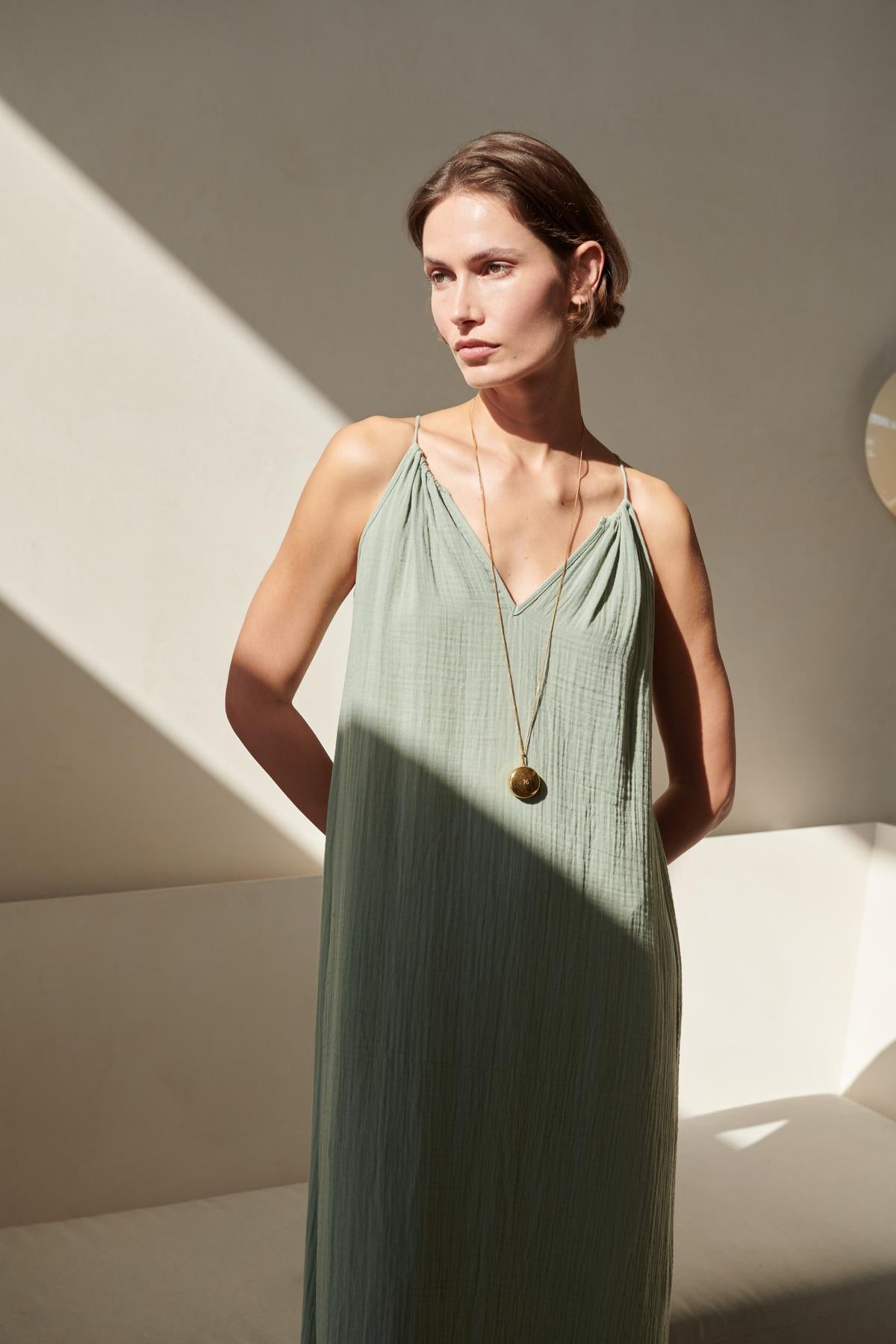   A woman in a Velvet by Jenny Graham CARRILLO DRESS stands in a sunlit room with shadows cast by a window, looking thoughtfully to the side. 