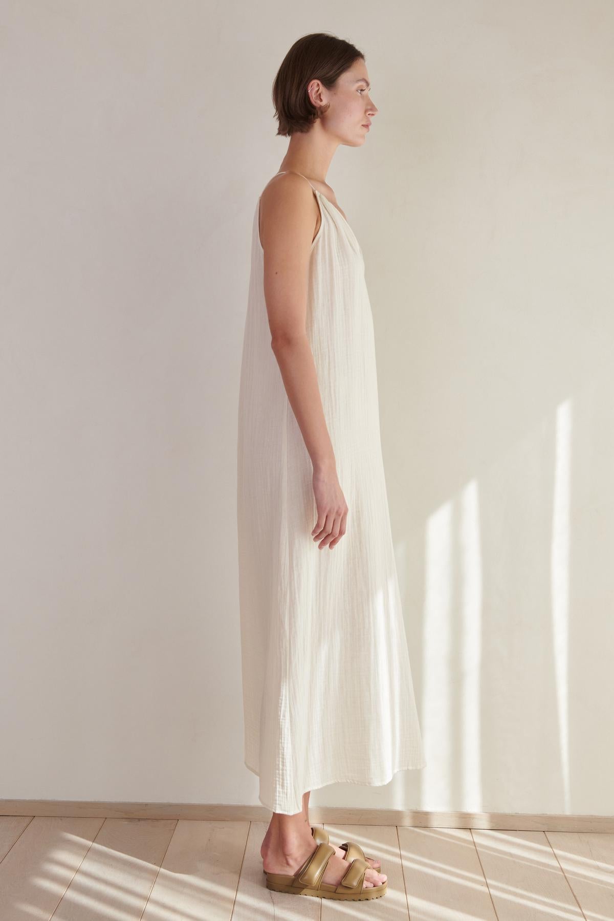   Woman standing in a sunlit room wearing a CARRILLO DRESS by Velvet by Jenny Graham with a V-neckline and pale green sandals, looking to the side. 