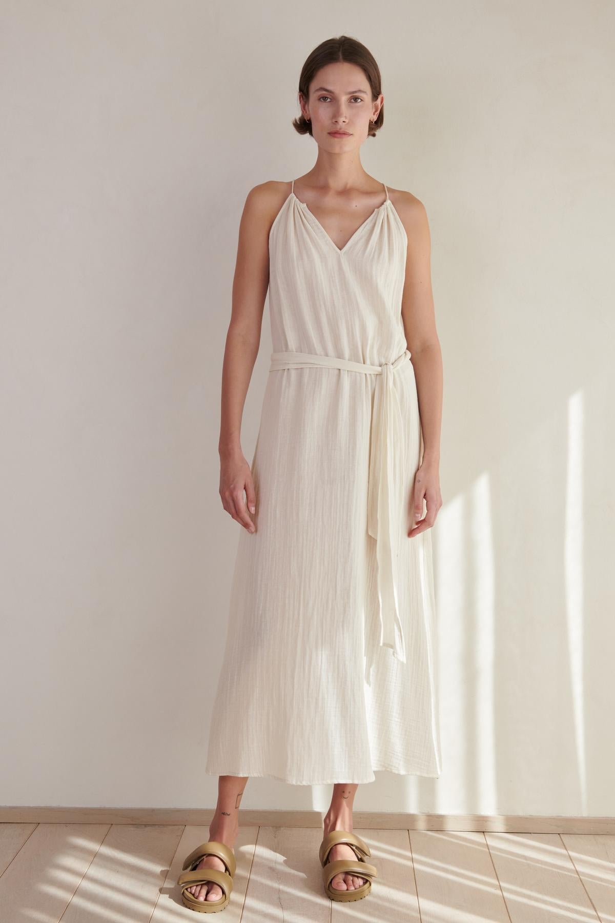   A woman in a white sleeveless CARRILLO DRESS by Velvet by Jenny Graham and gold sandals stands in a sunlit room with a neutral background. 