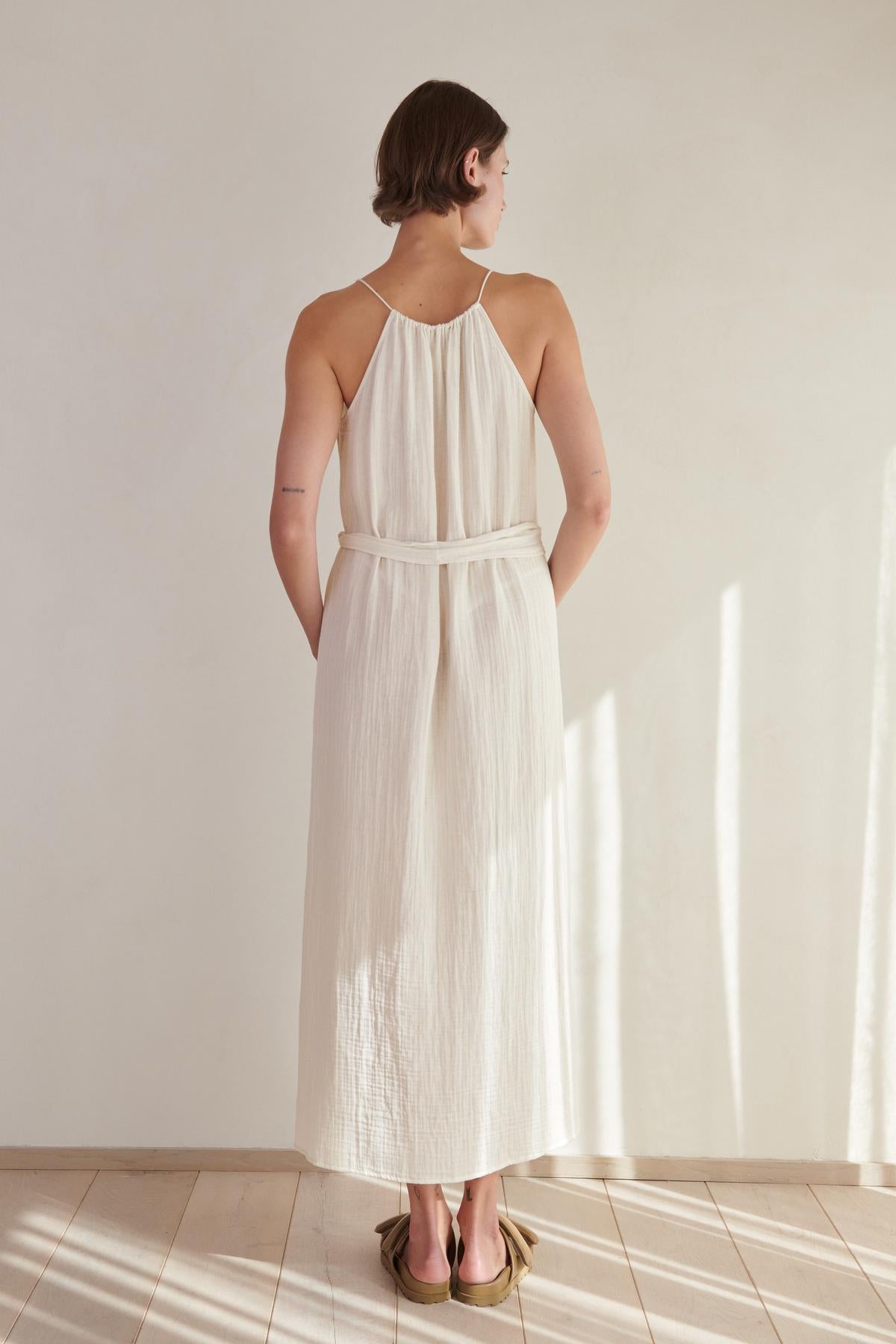   Woman in an elegant Velvet by Jenny Graham Carrillo Dress, a white cotton gauze halter-neck dress, standing with her back to the camera in a sunlit room. 