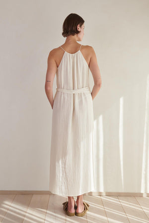 Woman in an elegant Velvet by Jenny Graham Carrillo Dress, a white cotton gauze halter-neck dress, standing with her back to the camera in a sunlit room.