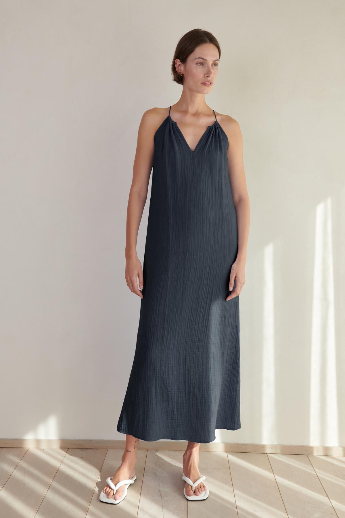   A woman in a Velvet by Jenny Graham CARRILLO DRESS dress and white sandals stands in a sunlit room with a tiled floor. 