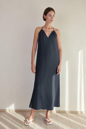 A woman in a Velvet by Jenny Graham CARRILLO DRESS dress and white sandals stands in a sunlit room with a tiled floor.