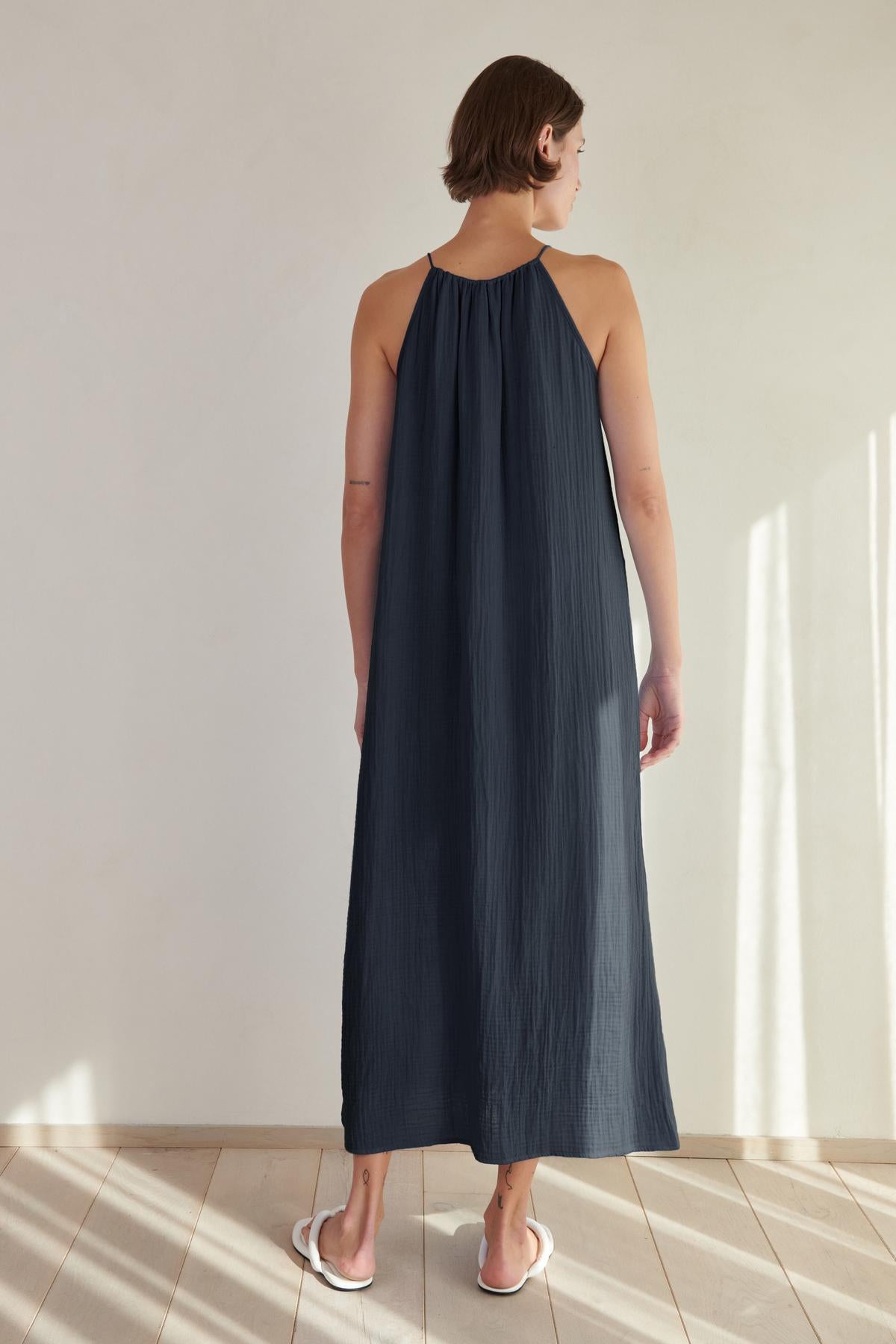   A woman in a navy blue cotton gauze Carrillo dress by Velvet by Jenny Graham standing in a sunlit room facing away from the camera. 