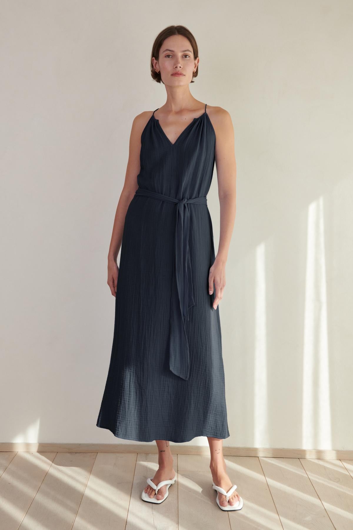   A woman in a navy blue sleeveless Carrillo dress by Velvet by Jenny Graham and white slides, standing in a softly lit room with shadows on the wall. 