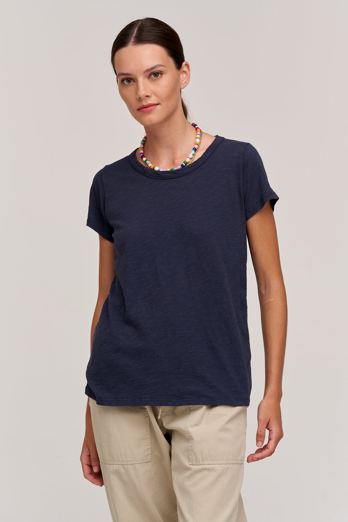   A person with straight hair is wearing a perfect navy TILLY TEE by Velvet by Graham & Spencer, beige pants, and a colorful beaded necklace. They are standing against a neutral background. 