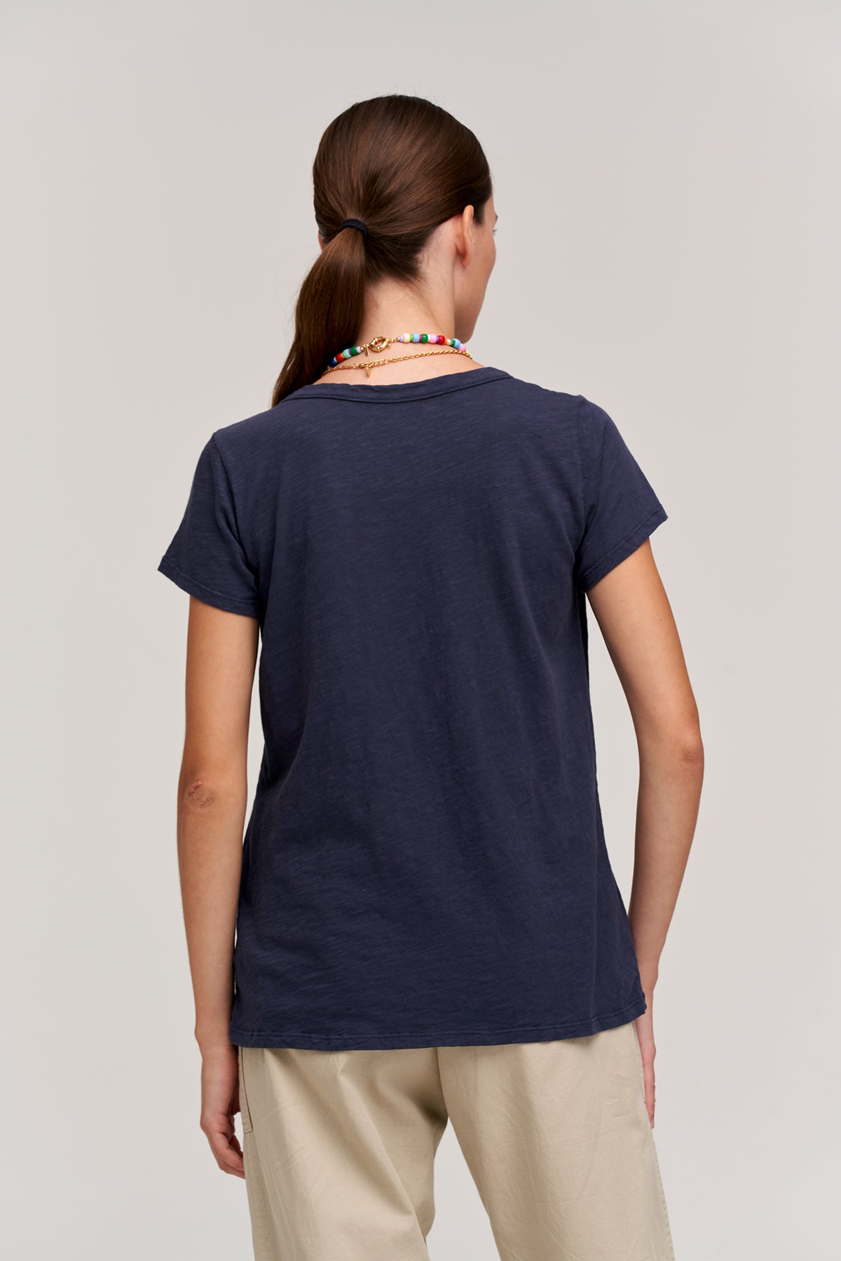   A person with a ponytail, wearing a navy blue TILLY TEE by Velvet by Graham & Spencer and beige pants, is seen from behind against a neutral background. They also have a colorful beaded necklace. 