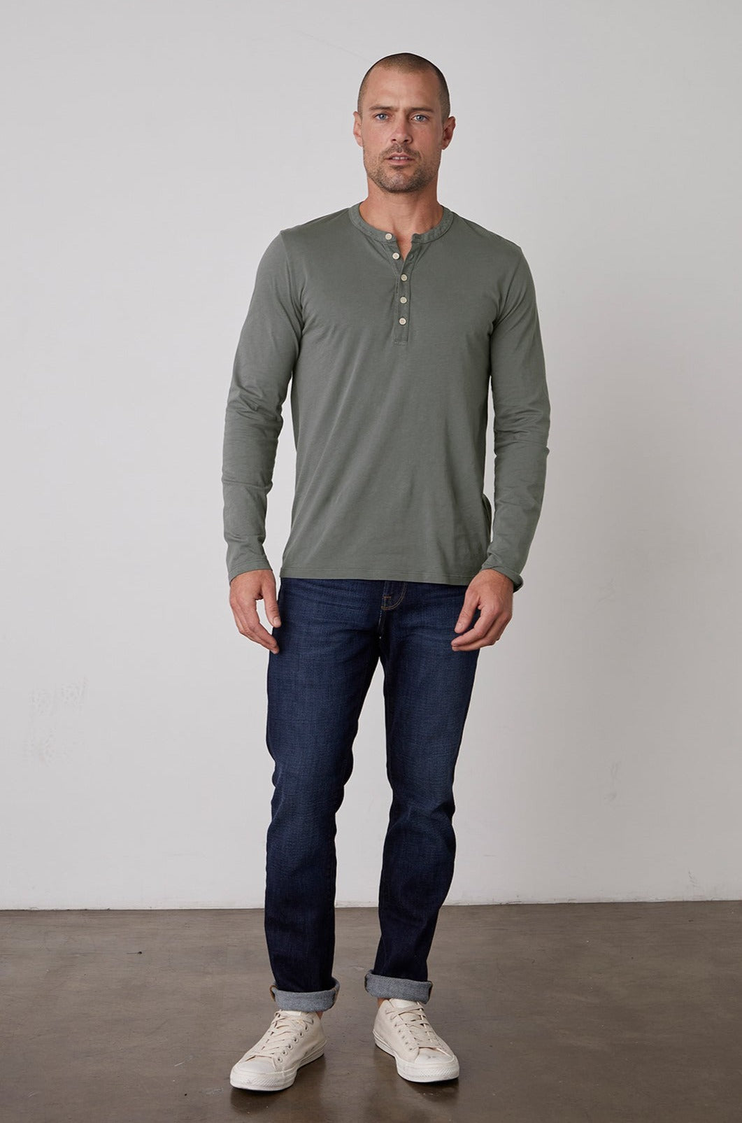 A man standing against a plain background is wearing a green ALVARO HENLEY by Velvet by Graham & Spencer, dark blue jeans, and white sneakers.-23778972467393