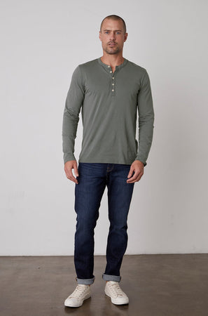 A man standing against a plain background is wearing a green ALVARO HENLEY by Velvet by Graham & Spencer, dark blue jeans, and white sneakers.