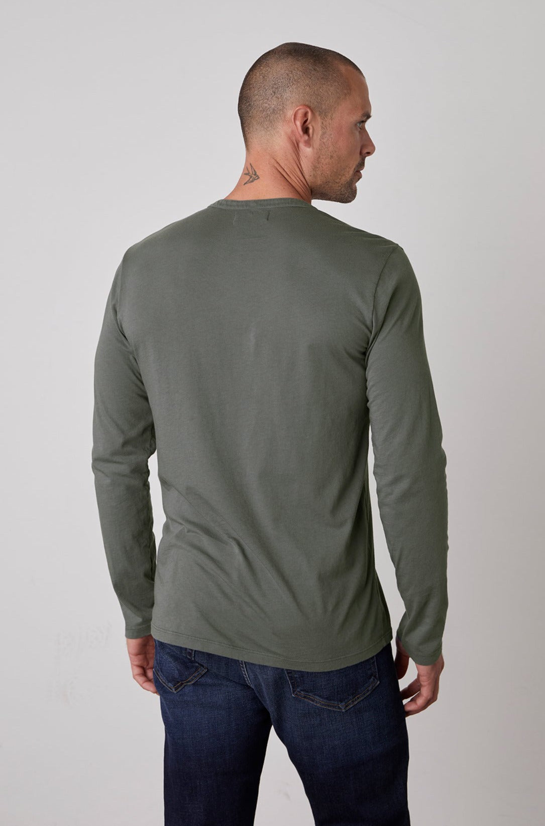 A person with a shaved head and a small neck tattoo is wearing a lightweight, long-sleeved green ALVARO HENLEY by Velvet by Graham & Spencer and blue jeans, viewed from the back.-23778972565697