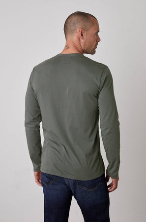A person with a shaved head and a small neck tattoo is wearing a lightweight, long-sleeved green ALVARO HENLEY by Velvet by Graham & Spencer and blue jeans, viewed from the back.