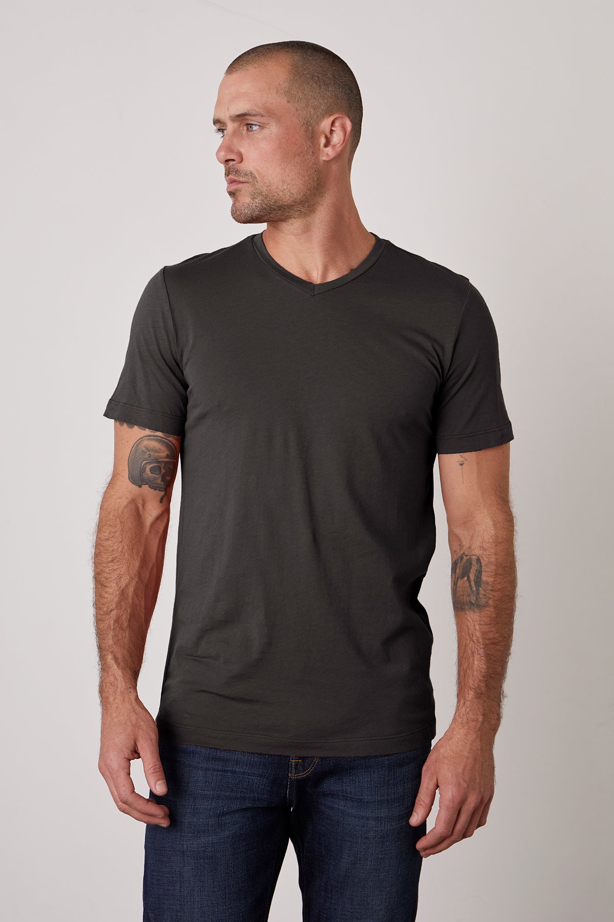   A man with a shaved head and tattoos on his forearms is wearing a plain, fitted black SAMSEN TEE by Velvet by Graham & Spencer and dark jeans. He is looking off to the side against a plain background. 