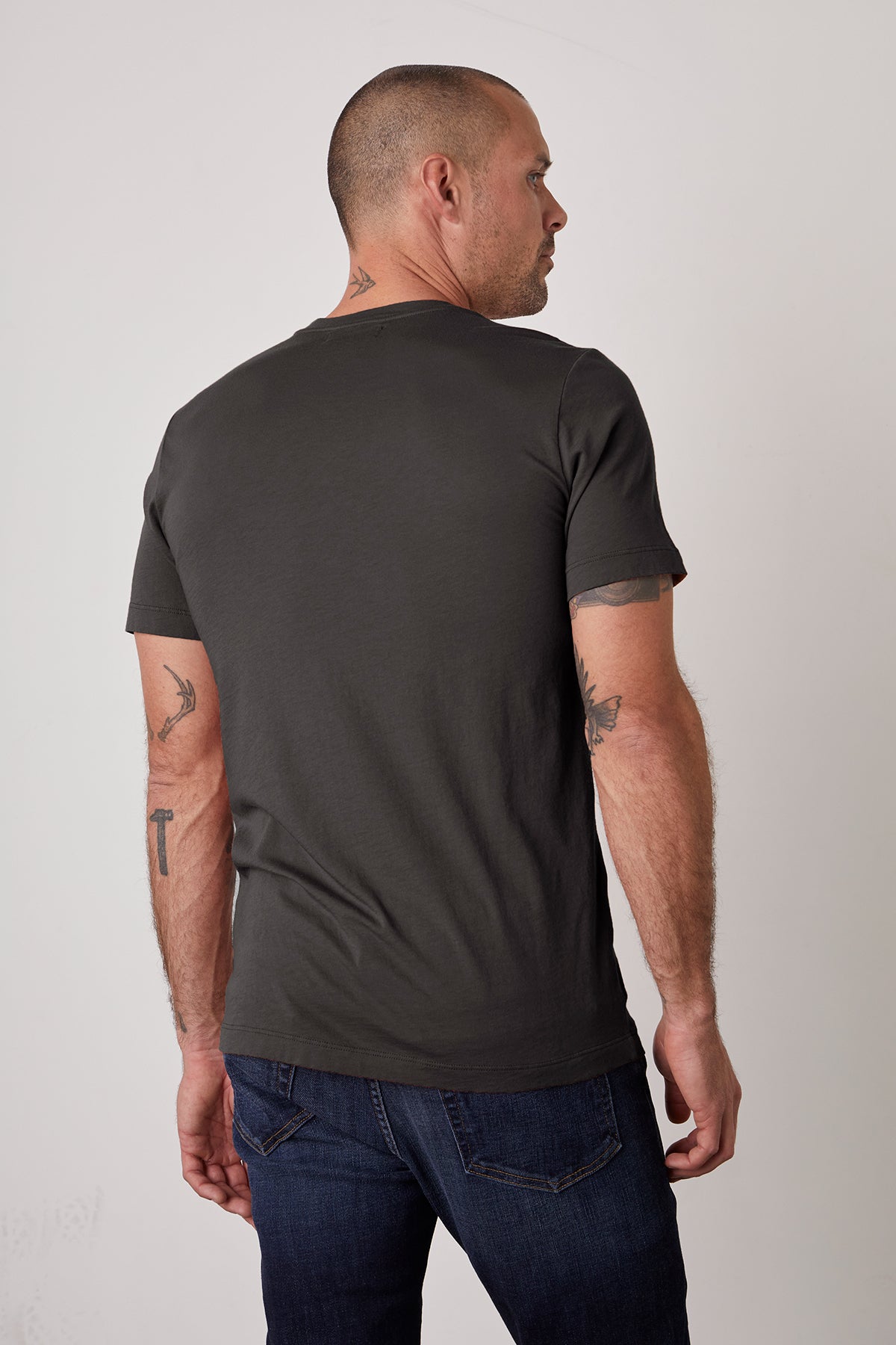   A man with a shaved head and tattoos on his neck and arms is standing and facing away, wearing a dark grey SAMSEN TEE by Velvet by Graham & Spencer that showcases a flawless fit and dark blue jeans. 