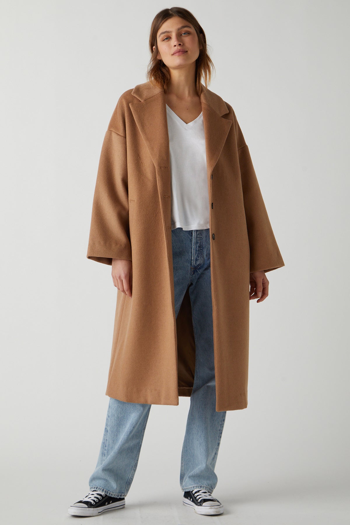   A person stands wearing a classic HUNTINGTON COAT by Velvet by Jenny Graham over a white V-neck top, blue jeans, and black and white sneakers. The chic essential outfit is set against a plain, light-colored background. 