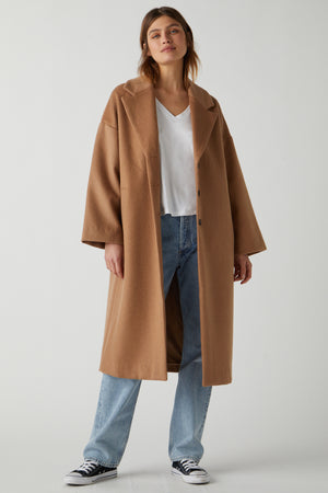 A person stands wearing a classic HUNTINGTON COAT by Velvet by Jenny Graham over a white V-neck top, blue jeans, and black and white sneakers. The chic essential outfit is set against a plain, light-colored background.