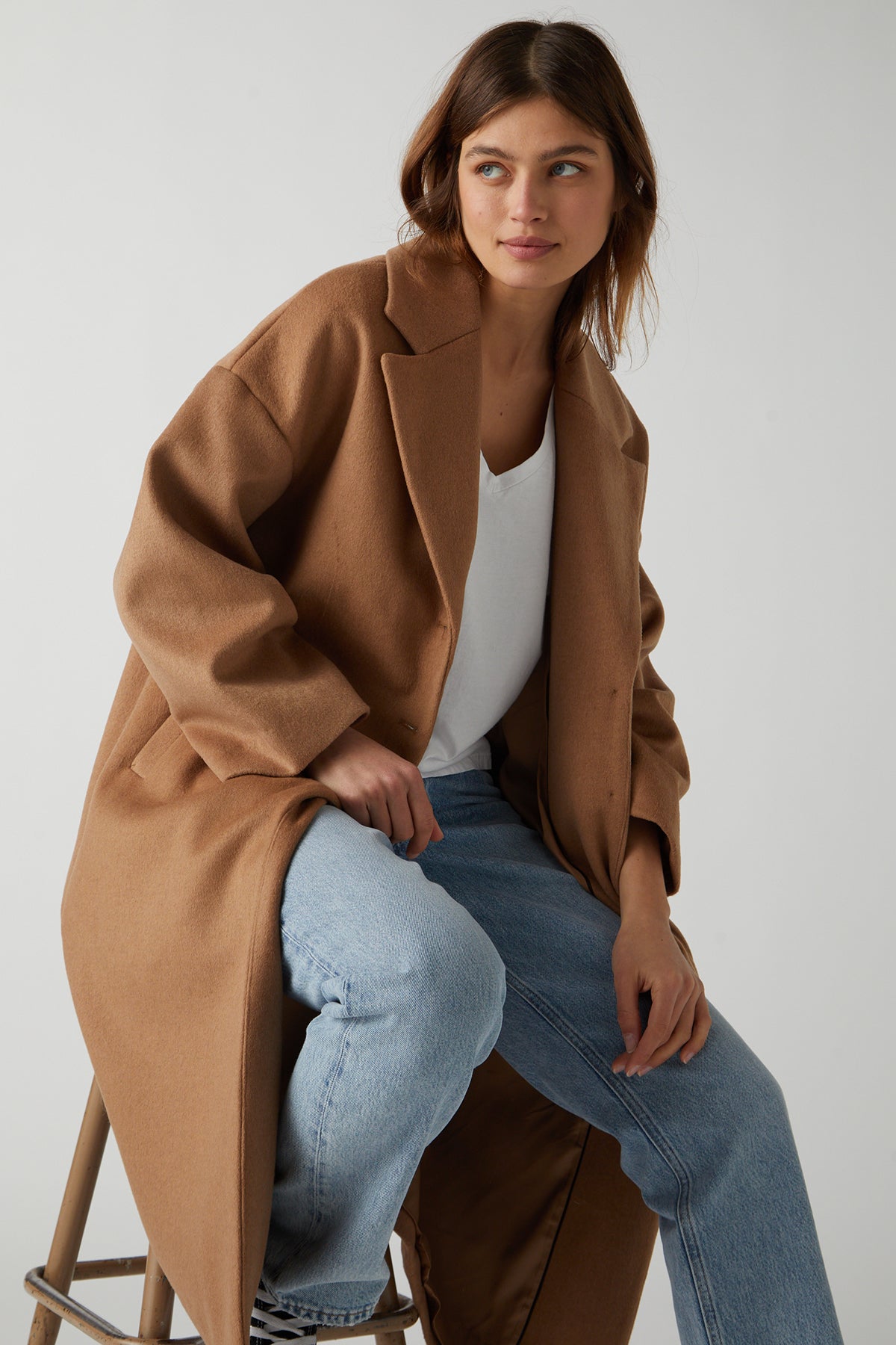   A person wearing a Velvet by Jenny Graham HUNTINGTON COAT over a white shirt and blue jeans, sitting on a stool and looking to the side. 