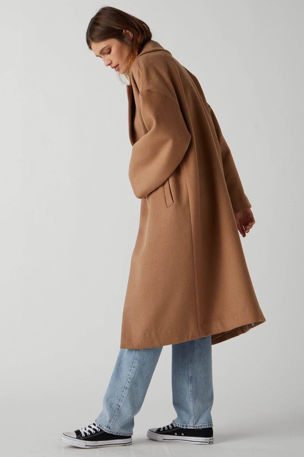   A person with shoulder-length hair stands in a neutral pose, wearing a chic essential, HUNTINGTON COAT by Velvet by Jenny Graham, in a classic camel color and soft wool blend. They pair it with light blue jeans and black sneakers against a plain background. 