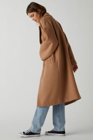 A person with shoulder-length hair stands in a neutral pose, wearing a chic essential, HUNTINGTON COAT by Velvet by Jenny Graham, in a classic camel color and soft wool blend. They pair it with light blue jeans and black sneakers against a plain background.