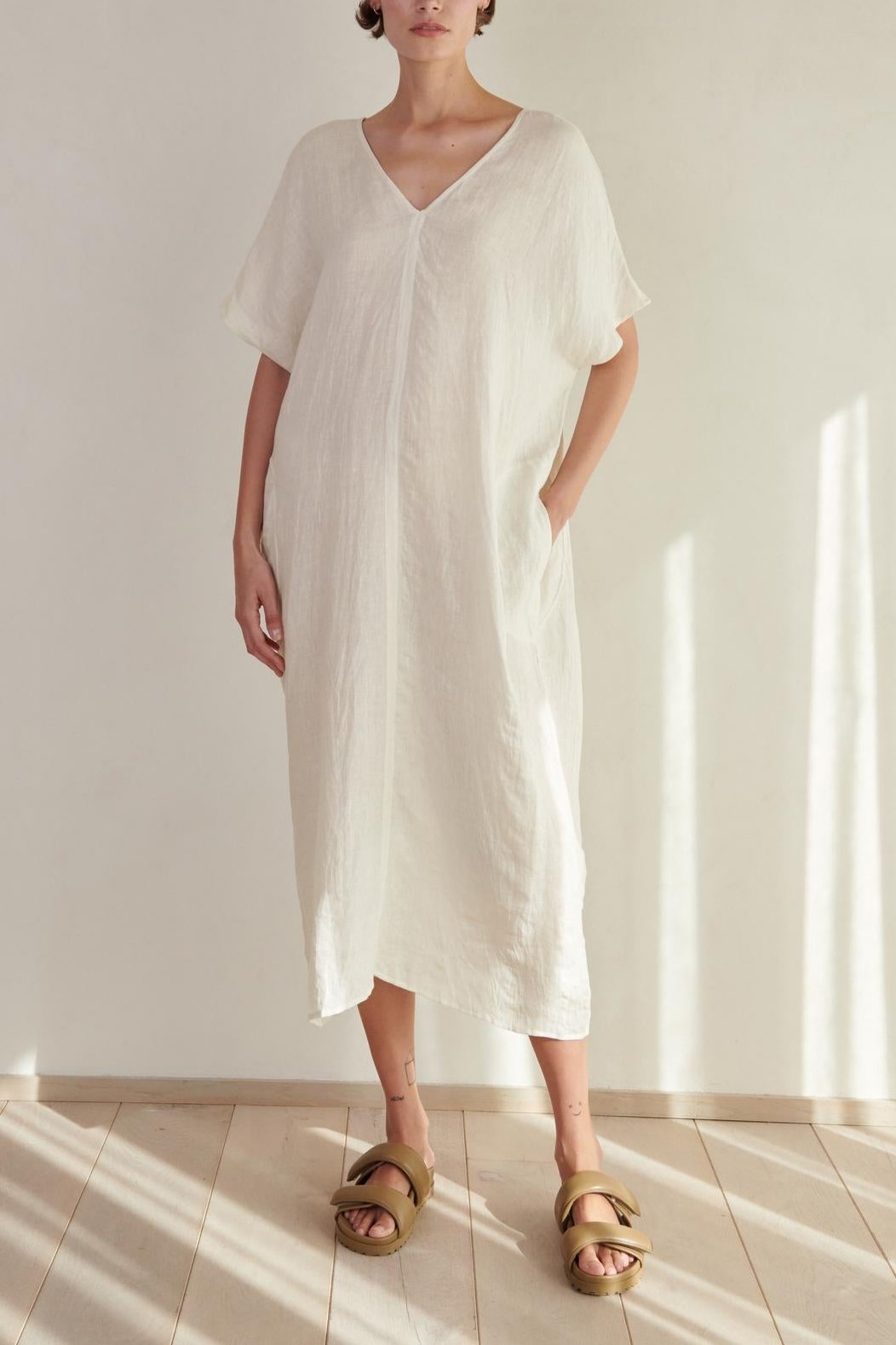   A woman wearing a long, white MONTANA LINEN DRESS with a V neckline and gold sandals stands in a softly lit room with a wooden floor. (Brand Name: Velvet by Jenny Graham) 