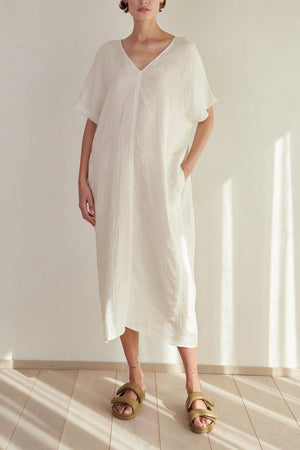 A woman wearing a long, white MONTANA LINEN DRESS with a V neckline and gold sandals stands in a softly lit room with a wooden floor. (Brand Name: Velvet by Jenny Graham)