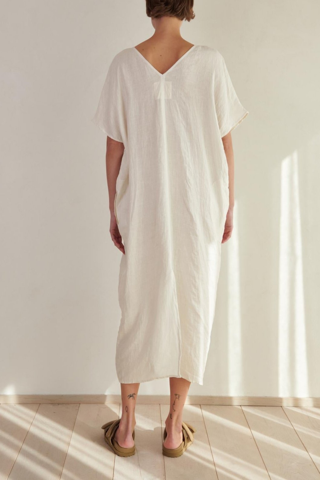 A woman wearing a Velvet by Jenny Graham Montana Linen Dress stands with her back to the camera in a bright, sunlit room.-26293194948801