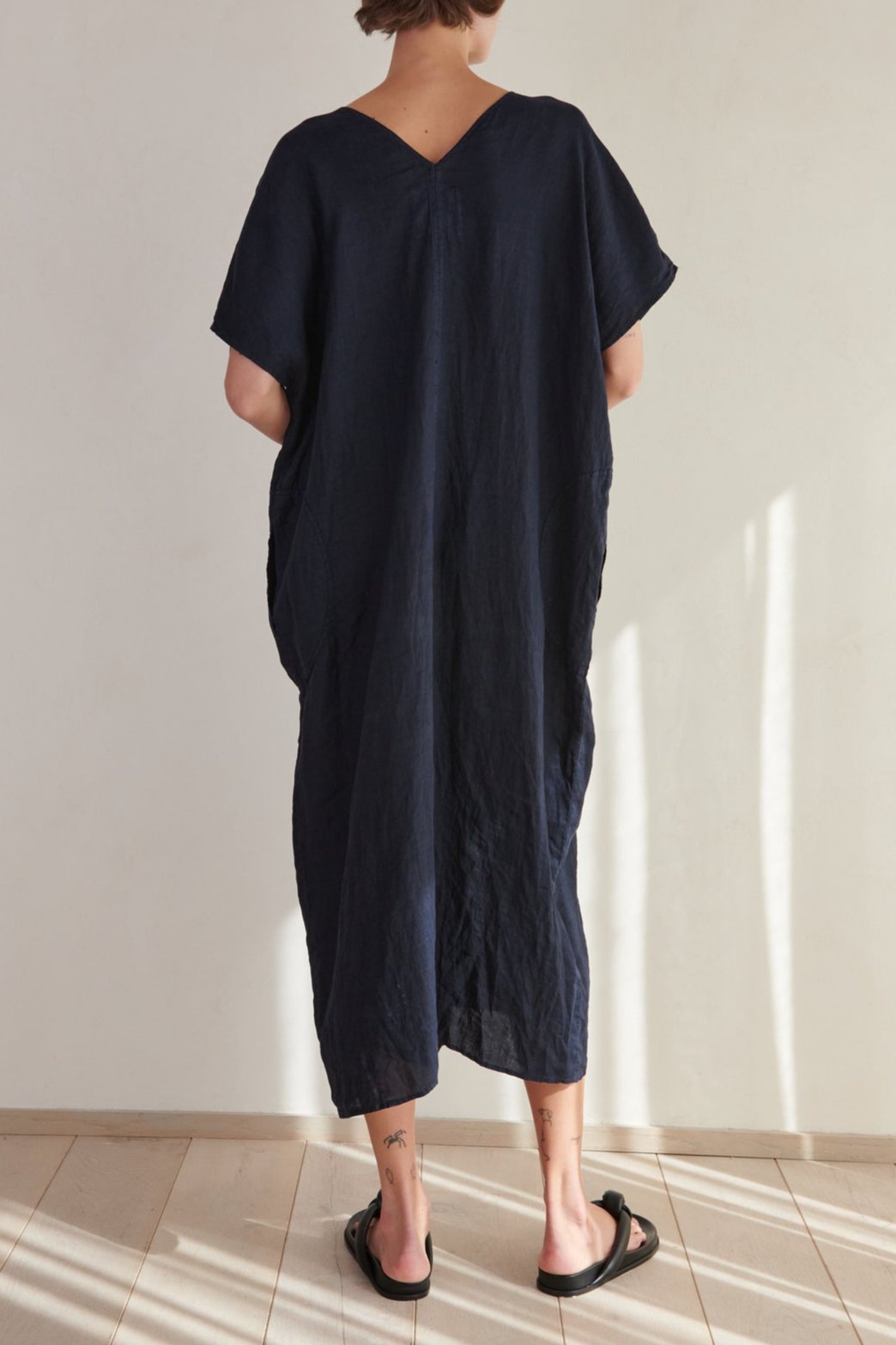   A woman in a Velvet by Jenny Graham Montana Linen Dress stands facing away from the camera, highlighting a loose fit and simple design, in a well-lit room with a neutral background. 